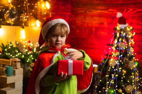 Lindo niño abriendo un regalo de Navidad. Niño alegre vestido de Papá Noel. Un niño con sombrero de Santa ayuda con el regalo de Navidad en una caja roja. — Foto de Stock