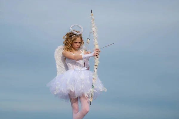 Little girl with angel wings and halo. Toddler girl wearing angel costume white dress and feather wings. Sweet angel girl. Portrait of little curly blond Angel girl. Concept of innocent child