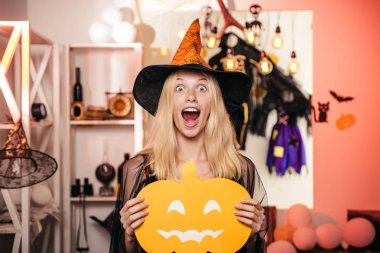 Beautiful young surprised woman in witches hat and costume holding pumpkin. Pumpkin head jack lantern. Trick or treat. Pretty young blond witch flying on the broom, isolated against orange background. clipart