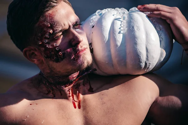 Muscular man with broken head. Close-up portrait of a man with zombie makeup. Scary and bloody muscular zombie man. Young handsome man with muscular body posing in street holding halloween pumpkin.