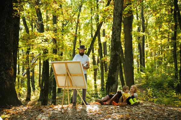 Painter with easel and canvas. Hipster artist with a beard in a hat creating art in the woods. Art concept. Painting in nature. Start new picture.