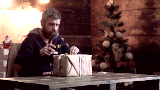 Diseño Cajas de regalo hechas a mano. Hombre barbudo envolviendo regalos de Navidad en papel. Retrato de un brutal Santa Claus maduro. Hombre barbudo en jersey navideño. Casa Ambiente de Navidad . — Vídeos de Stock