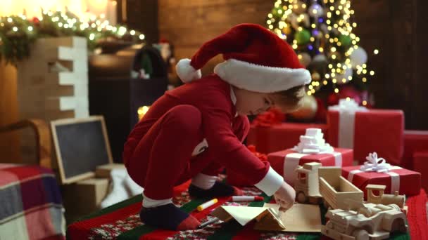 Lycklig söt barn i Santa hatt med nuvarande har en jul. Kid ha kul nära julgran inomhus. Happy Child med julklapp ask. Barn med en julklapp på trähus bakgrund. — Stockvideo