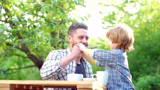 小男孩和英俊的父亲在花园里吃早餐。为人 父母。家庭食物和饮料。快乐的父亲与他的儿子坐在桌子上吃健康的食物与滑稽的表情在脸上. — 图库视频影像