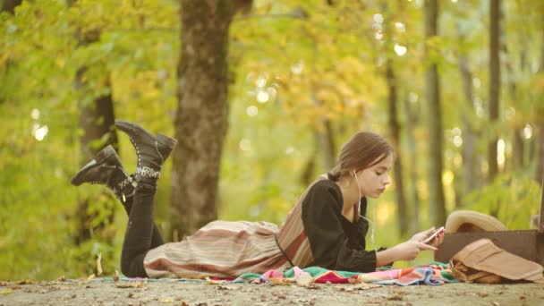 幸せな女性ヒップスターは、秋の背景にスマートフォンで音楽を聴きます。公園の女性モデル。秋の背景の上に楽しみを持っている若い女性. — ストック動画