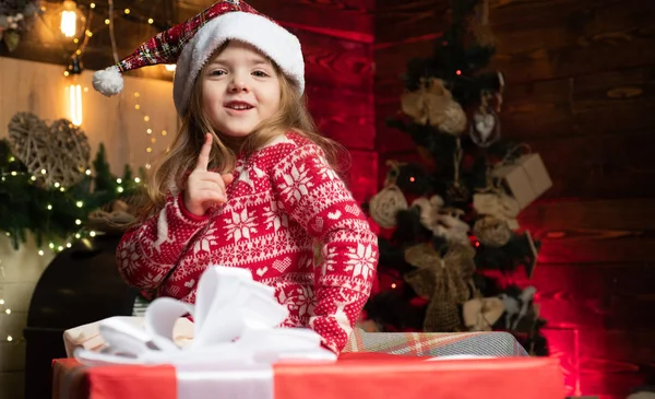 Porträtt liten flicka med gåva på trä jul bakgrund. Söt liten flicka som kommer till julgran och tittar på hennes nya år gåva. Barnens slitage trender i vinter 2020. — Stockfoto