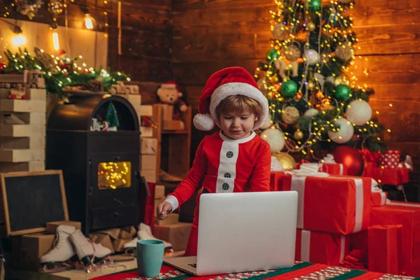 Pequeño genio. Santa ayudante. Niño Santa sombrero y traje de divertirse. Niño con portátil cerca del árbol de Navidad. Comprar regalos de Navidad en línea. Concepto de compras navideñas. Servicio de regalos —  Fotos de Stock