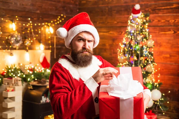 Hipster in red Santa hat holding present. Styling Santa hipster with a long beard posing on the Christmas wooden background. Winter emotion. — Stock Photo, Image