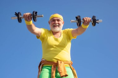 Yaşlı adam ağırlıkları kaldırıyor. Spor salonunda ağırlıklarla çalışan kıdemli adam. Mutlu kıdemli adam kamera bakıyor dumbbell ile. Mavi gökyüzü arka üzerinde büyükbaba sporcu portre.