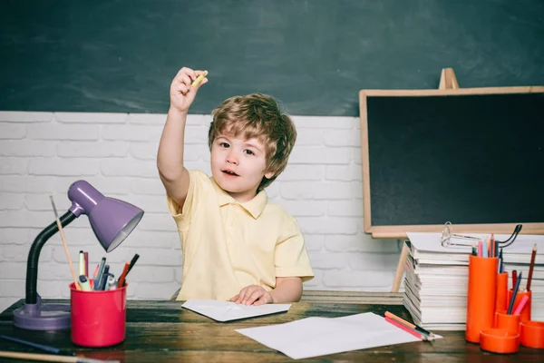 Allievo bambino con espressione faccia felice vicino alla scrivania con forniture scolastiche. Il ragazzo si prepara per andare a scuola. Allegro sorridente piccolo allievo divertirsi contro lavagna . — Foto Stock