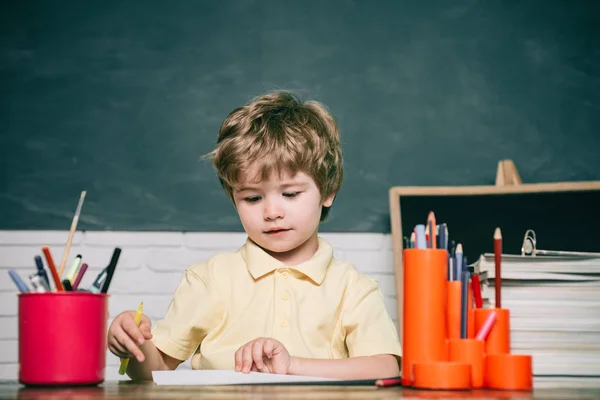 Barn från grundskolan med bok och väska. Barnet är redo att svara med en svart tavla på en bakgrund. Happy Cute flitig barn sitter vid ett skrivbord inomhus. — Stockfoto