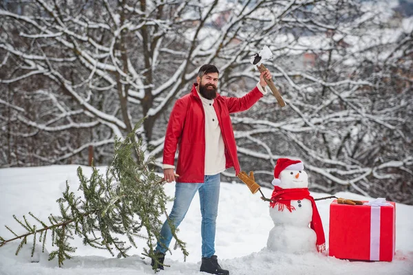 Un beau jeune homme avec un bonhomme de neige porte un sapin de Noël. Joyeux Noël et bonne année carte de voeux. Bonhomme de neige avec cadeau . — Photo