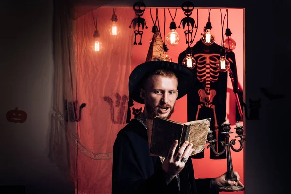 Hipster aterrador con barba en sombrero de bruja y camisa a cuadros. Feliz hipster con barba ensangrentada. Brujo malvado con el pelo rojo y una barba en una capa negra leyendo un libro de hechizos. concepto de gente de Halloween . —  Fotos de Stock