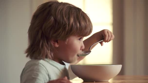 Retrato de un dulce niño riéndose con el pelo rubio comiendo de un plato sosteniendo la cuchara de cerca. Niño sentado en la mesa y comiendo bocadillos de leche. Infancia . — Vídeos de Stock