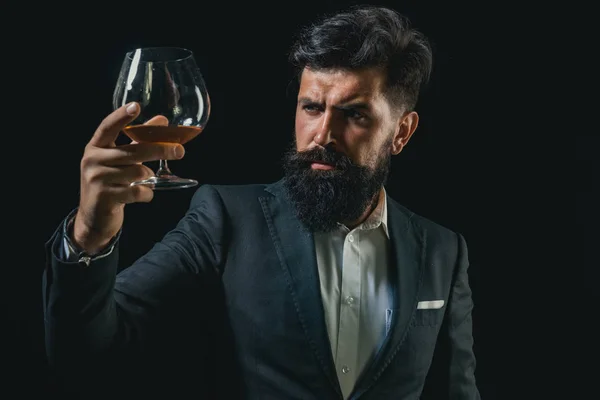 Degustacja elitarnego alkoholu. Pewnie Brodaty człowieka w czarnym kolorze z kieliszek koniaku. Człowiek z brodą trzyma szklankę brandy. — Zdjęcie stockowe