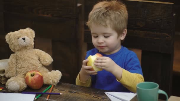 Söt blond barn pojke sitter i trä stol och äta stort rött äpple på trä bakgrund. Skol uppehåll. Hungrig unge bita äpple i klassrummet. Liten pojke leker med papper planet och nallebjörn. — Stockvideo