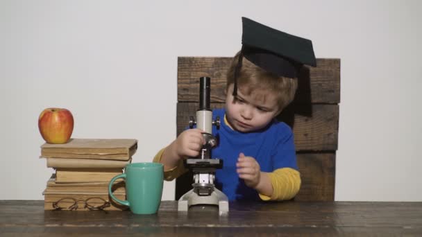 Concept de gamin intelligent. Enfant avec une expression faciale grave tient le microscope. Premier intéressé par l'étude, l'apprentissage, l'éducation. Le gamin en bonnet académique travaille au microscope. Concept de génie . — Video