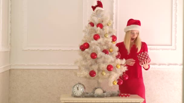 Santa mujer sosteniendo la caja de regalo de Navidad en la pared blanca. Una mujer sensual. Retrato de moda de chica modelo en el interior con árbol de Navidad . — Vídeos de Stock