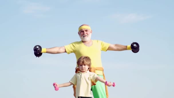 Senior man och barn i Family Health Club. Far och son spotting. Senior Fitness man utbildning med hantlar på blå bakgrund. Farfar och barn i gymmet tränar med vikter. — Stockvideo