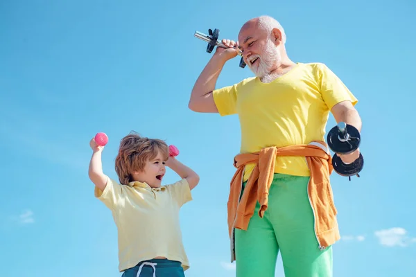 Hombre mayor y niño ejercitándose en el cielo azul. Salud estilo de vida alegre. Retrato de un padre y un hijo sanos haciendo ejercicio con pesas sobre el fondo azul del cielo. — Foto de Stock