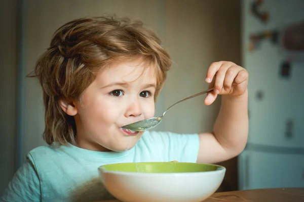 Śmiech Cute dziecko chłopiec siedzi w krzesełko i jedzenie na niewyraźne tło. Wesołe dziecko zjada jedzenie z łyżką. Rodzicielstwa. Mały chłopak siedzi przy stole i jedzenia mleka przekąskę. — Zdjęcie stockowe