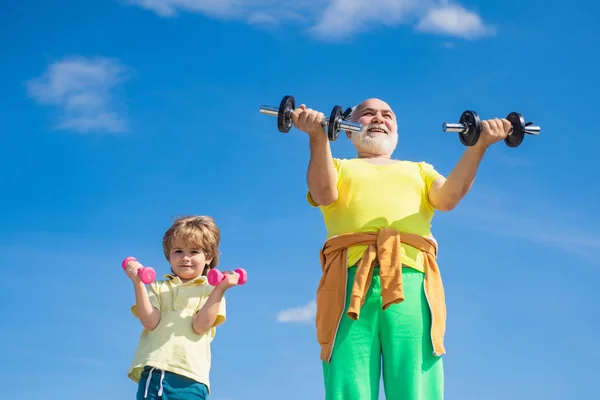 Apa és fia pecsételő. Apa és fia épít bicepsz és tricepsz. Portré egy egészséges apa és fia dolgoznak ki a súlyzók felett kék ég háttér. — Stock Fotó