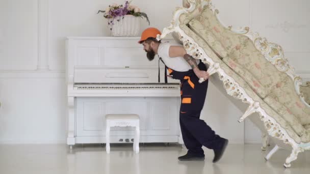 Concept de réinstallation. Courier livre des meubles en cas de déménagement, déménagement. Chargeur déplace canapé, canapé. Homme avec barbe, travailleur en salopette et casque porte canapé sur le dos, fond blanc . — Video