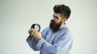 Sakallı ve bıyıklı hipster. Sıkılmış, uykulu yüzlü adam battaniyenin üzerine oturur, çalar saati tutar. Adam sabahları vakit harcıyor, beyaz arka plan. Tembel ve uyuyakalmış kavram. Adam alarmı kuruyor..