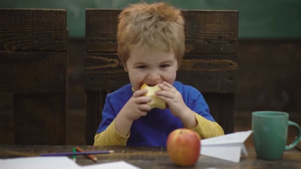 Lindo niño rubio sentado en una silla de madera y comiendo fruta de manzana roja grande. Retrato sobre fondo borroso, primer plano. Descanso escolar. Chico hambriento comiendo manzana en clase. Almuerzo escolar. Regreso a la escuela . — Vídeos de Stock