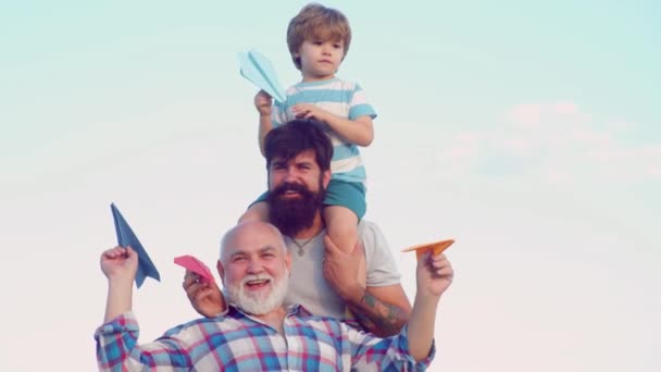 Ευτυχισμένο παιδί με τον πατέρα και τον παππού να παίζουν με το αεροπλάνο χαρτί παιχνίδι ενάντια στο καλοκαιρινό φόντο του ουρανού. Ο παππούς κάνει παιχνίδι. Χαριτωμένο γιο με τον μπαμπά και τον πατέρα που παίζουν υπαίθρια. — Αρχείο Βίντεο