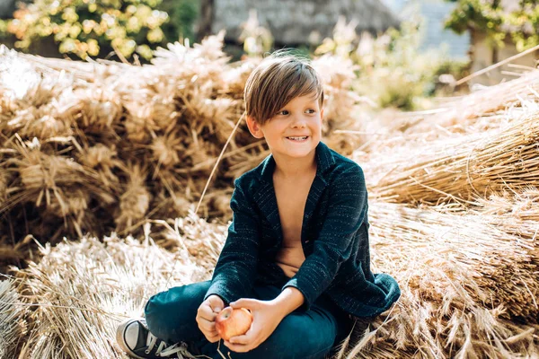 Fair-haired boy lies on hay background and eats an apple. Sale for entire autumn collection, incredible discounts and wonderful choice. Bye summer - hi autumn. Back to school.