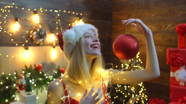 Karácsonyi party. Szexi szőke nő tartja piros karácsonyi labdát a karácsonyfa backgroung. Karácsonyi díszek. Új év party. — Stock videók