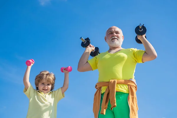 Aranyos gyerek és magas rangú ember gyakorlása a súlyzók. Boldog kis fiú és nagyapja emelési súlyzók kék ég háttér. Egészséges neveltetés. — Stock Fotó