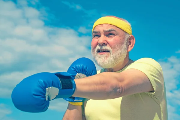 Um homem fixe a lutar. O melhor exercício cardiológico. Homem sénior a usar luvas de boxe. Retrato de um determinado boxeador sênior sobre fundo céu azul . — Fotografia de Stock