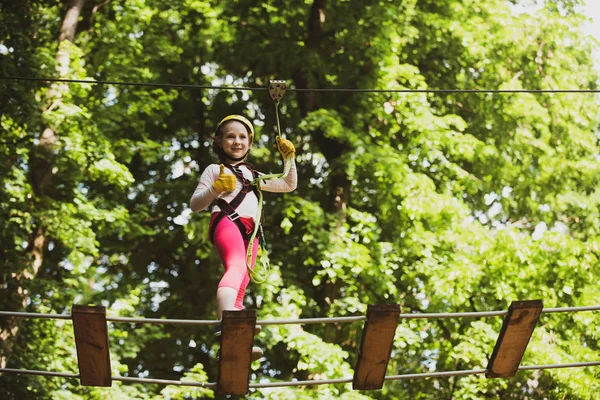 Eco Resort Activities. Little girl climbing on high rope park. Little girl concept. Cute little girl in climbing safety equipment in a tree house or in a rope park climbs the rope.