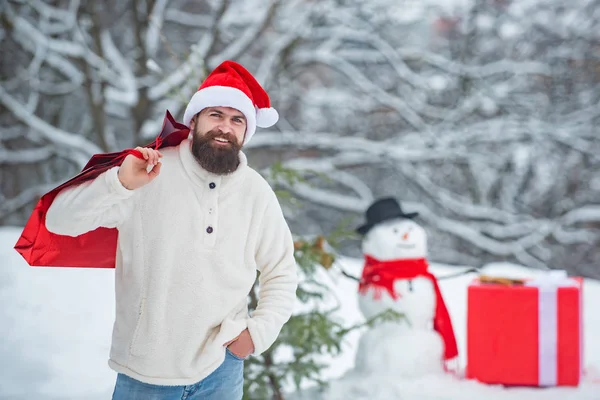 Homme avec cadeau rouge jouant avec bonhomme de neige dans le parc d'hiver. Homme en robe de Noël avec cadeau près du fond de Noël. Holly joyeux swag Noël et le nez . — Photo