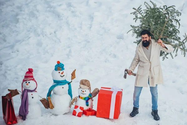 Man with beard bears home a Christmas tree. Winter emotion. Merry Christmas and Happy Holidays. Young lumberjack bears fir tree in the white snow background.