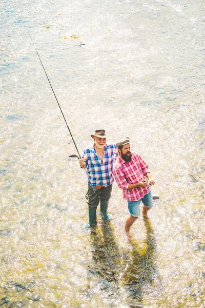 世代の年齢:祖父と父。川で釣り。若い男と老人は釣りを飛ぶ。川に息子を連れ、釣り竿を持って釣りを楽しむ父. — ストック写真