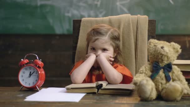 Malá holčička sedí u svého stolu s otevřenou knihou, medvídkem a poplašným hodinami, které si opřela hlavu o pozadí školní tabule. Zpátky do školy. Vzdělání, koncepce učení. Roztomilá holčička ve třídě — Stock video