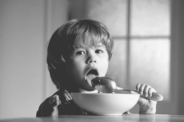 Ung unge som sitter på bordet och äter med lustiga ansiktsuttryck. Trevlig barnfrukost. Trevlig barnfrukost. Glada baby barn äter mat själv med sked. — Stockfoto