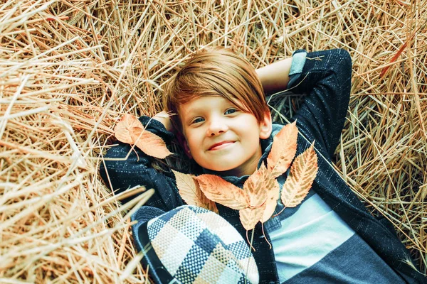 Leende liten pojke leker med blad och tittar på kameran. Kid boy ligger på hö. Söt liten barn pojke håller bladguld på gården byn bakgrund. — Stockfoto