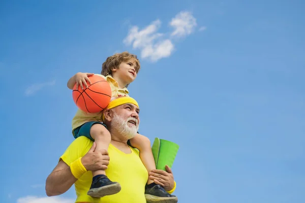 Preparándose para el entrenamiento matutino en el parque. Feliz niño sonriente en el hombro abuelo mirando a la cámara. Niño se sienta sobre los hombros de su abuelo. — Foto de Stock