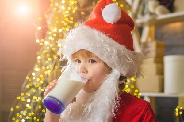 Happy New Year. Santa Claus. Happy kids Santa Claus with glass of milk and cookie. Cheerful little Santa Claus holding glass with milk and cookie with fireplace and Christmas Tree in the background. — Zdjęcie stockowe