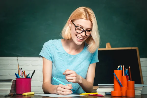 Glücklich beiläufige Studentin mit über Tafel Hintergrund. Eine junge Frau sitzt am Tisch und erledigt Aufgaben. Studentin denkt über Studium nach. — Stockfoto