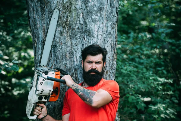 Woodcutter with axe or chainsaw in the summer forest. Deforestation. Handsome young man with axe near forest. The Lumberjack working in a forest. Chainsaw. — Stock fotografie