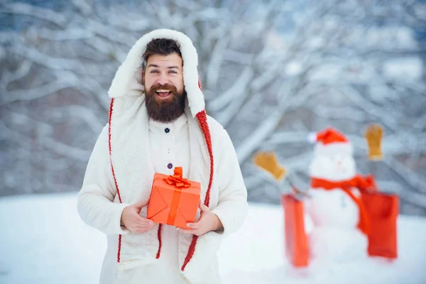 Biglietto di auguri natalizio. Ritratto invernale di hipster bello in neve Giardino con regalo fanno pupazzo di neve. Uomo con regalo rosso che gioca con pupazzo di neve nel parco invernale. — Foto Stock