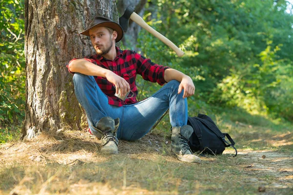 Trabalhador lenhador sentado na floresta. Lumberjack com machado no fundo da floresta. Homem a fazer trabalho de homem. Trabalhador lenhador de pé na floresta com machado. Descanso após trabalho duro . — Fotografia de Stock