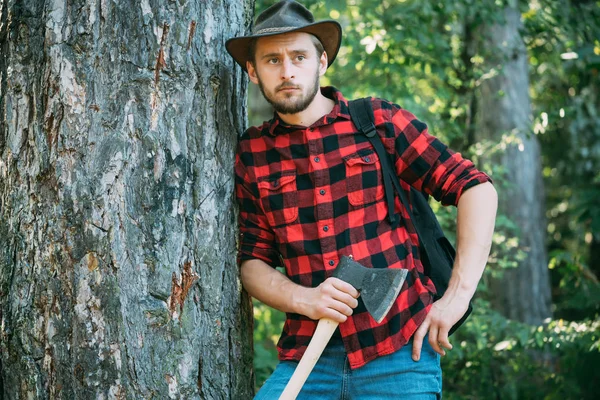 Knappe man met bijl. Handsome man houthakker met een grote bijl onderzoekt de boom voor het kappen. Sterke man houthakker met een bijl in een geruit shirt. — Stockfoto