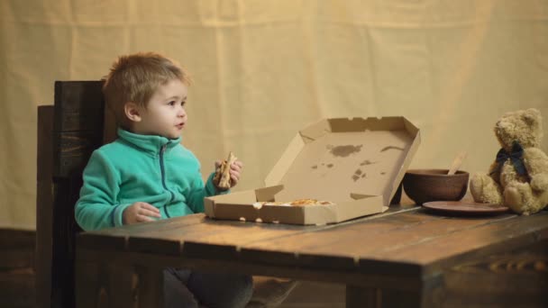 Chłopiec jedzenia pizzy siedzi na drewnianym stole z niedźwiedziem zabawka. Zabawny chłopak i Miś jedzą pizzę na tle juta. Smaczne pizze. Tło burlap. Foodów. Chłopiec z niedźwiedziem i pizzą. Niezdrowe jedzenie. — Wideo stockowe