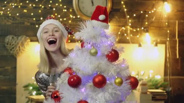 Chica alegre mira desde el árbol de Navidad. Chica de Navidad. Mujer con copa de champán y sombrero rojo de Navidad escondido detrás del árbol de Navidad. Feliz Navidad y Feliz Año Nuevo. Chica divertida de Año Nuevo . — Vídeos de Stock